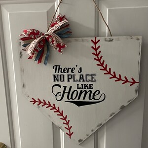 May include: A baseball-themed wooden home plate sign. The white sign features red stitching, a decorative bow, and the phrase "There's No Place Like Home" in black lettering. A jute rope is used for hanging.