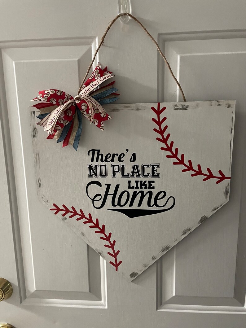 May include: A baseball-themed wooden home plate sign. The white sign features red stitching, a decorative bow, and the phrase "There's No Place Like Home" in black lettering. A jute rope is used for hanging.