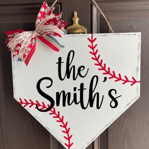 May include: White baseball home plate-shaped sign with the text "the Smith's" in black script. Red stitching detail mimics a baseball. A decorative bow with baseball-themed ribbon and a jute hanger complete the design.