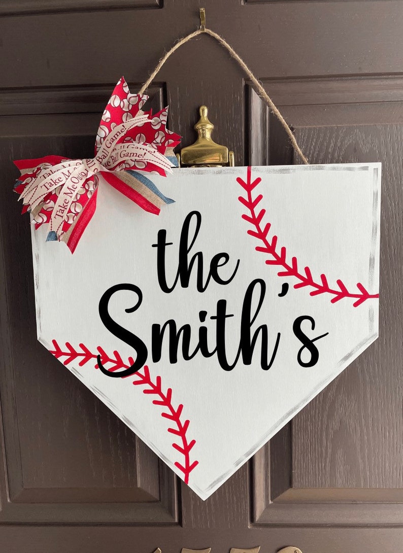 May include: White baseball home plate-shaped sign with the text "the Smith's" in black script. Red stitching detail mimics a baseball. A decorative bow with baseball-themed ribbon and a jute hanger complete the design.