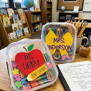 May include: Two clear zippered pouches filled with school supplies. One pouch features a red apple decal labeled 'MS. LIBBY 2ND GRADE' with a pencil graphic. The other features a yellow bow decal labeled 'MRS. ANDERSON' with various pens inside.