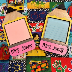 May include: Two pencil-shaped memo holders with pink and blue notepads. The holders are yellow and wood-toned with black accents and the name "Mrs. Jones" in black script. The background is a colorful school-themed fabric.