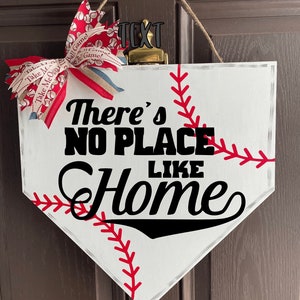 May include: A baseball-themed home decor sign shaped like home plate. The white sign features red stitching, the phrase "There's NO PLACE LIKE Home" in black, and a decorative bow with baseball and text patterns.