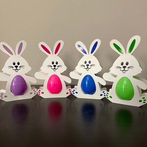 May include: Four white bunny-shaped Easter egg holders with different colored eggs. The bunnies are standing on a brown surface. The eggs are purple, pink, blue, and green.