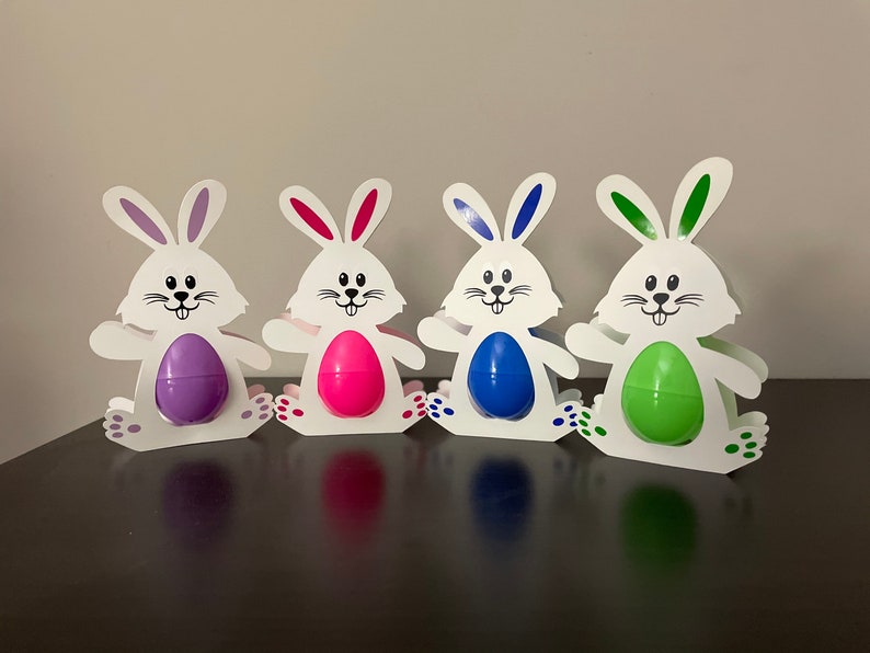 May include: Four white bunny-shaped Easter egg holders with different colored eggs. The bunnies are standing on a brown surface. The eggs are purple, pink, blue, and green.
