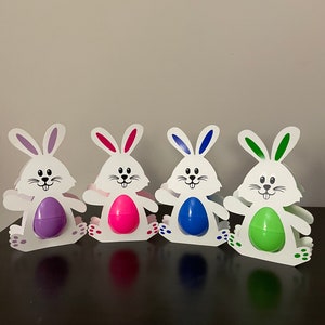 May include: Four white bunny-shaped Easter egg holders with different colored eggs. The eggs are purple, pink, blue, and green.