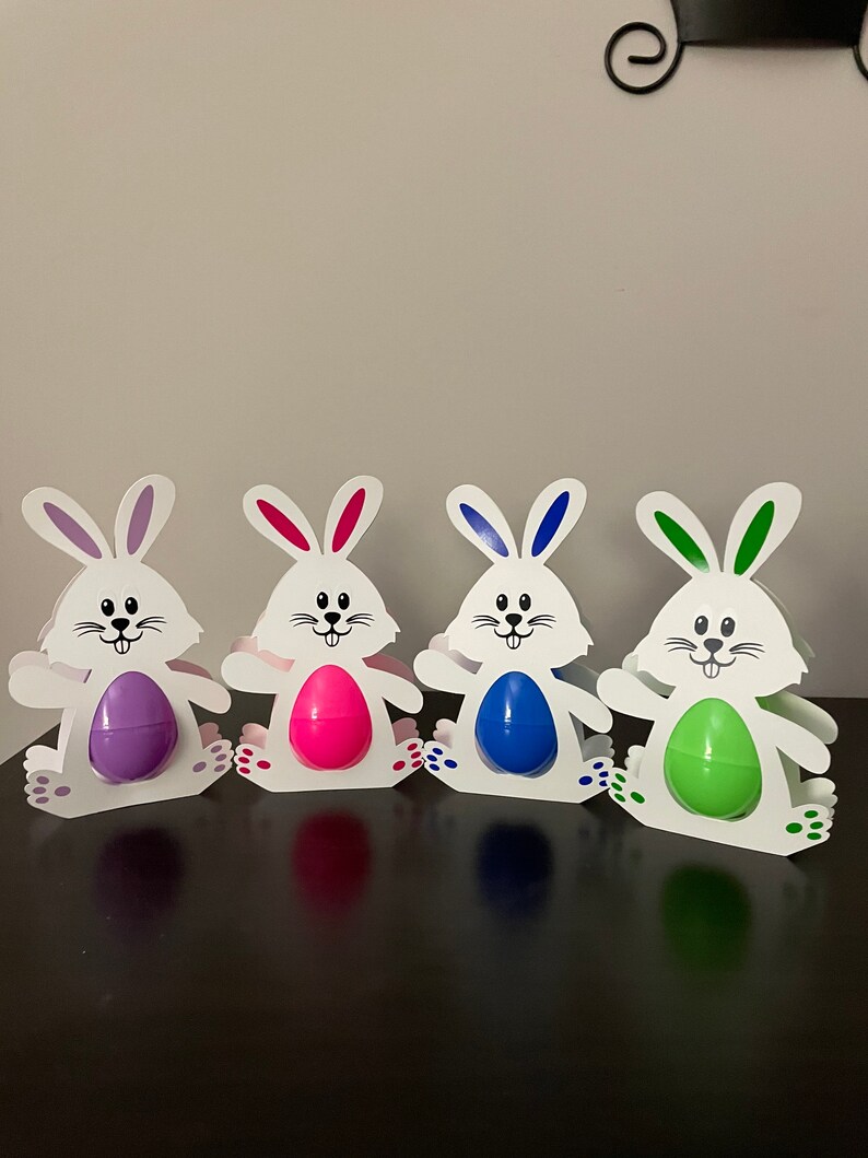May include: Four white bunny-shaped Easter egg holders with different colored eggs. The eggs are purple, pink, blue, and green.
