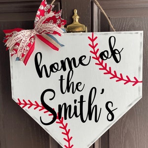 May include: White baseball home plate-shaped sign with the text "home of the Smith's" in black script. Red stitching detail and a red, white, and blue bow add a decorative touch. A jute rope is used for hanging.