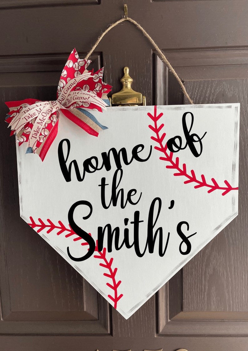 May include: White baseball home plate-shaped sign with the text "home of the Smith's" in black script. Red stitching detail and a red, white, and blue bow add a decorative touch. A jute rope is used for hanging.