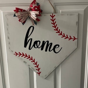 May include: White wooden home plate-shaped sign with red baseball stitching and the word "home" in black script. A decorative bow with baseball-themed fabric accents the top, hanging from a jute string.