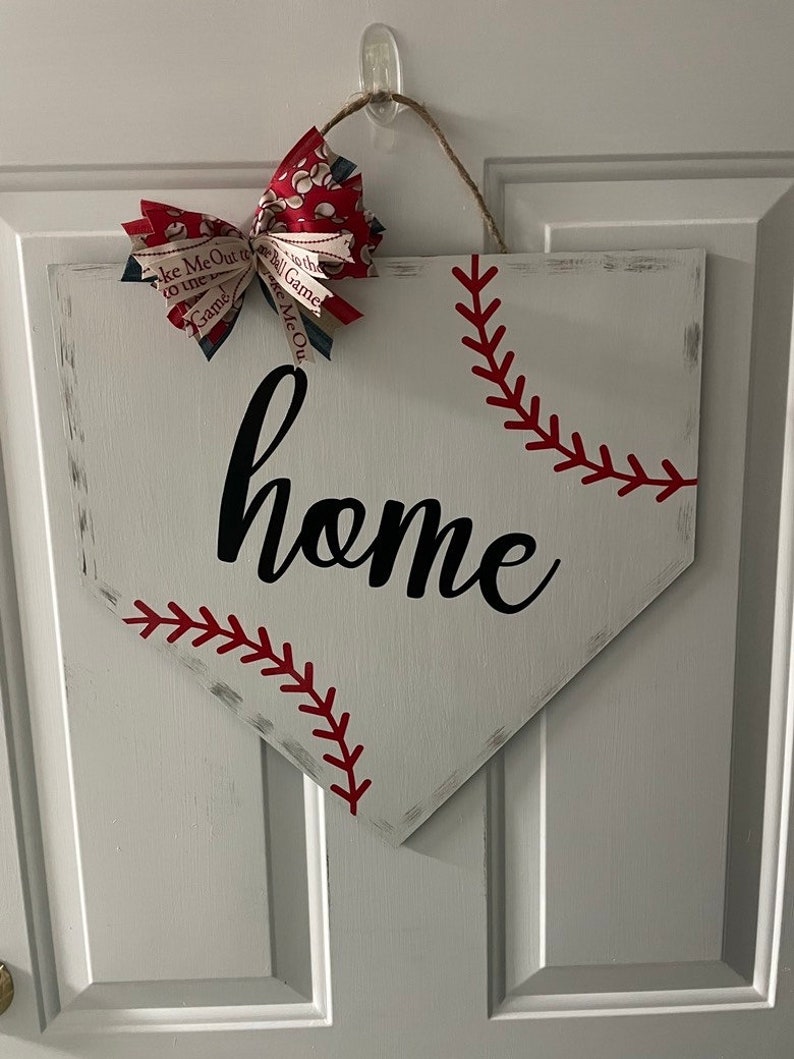 May include: White wooden home plate-shaped sign with red baseball stitching and the word "home" in black script. A decorative bow with baseball-themed fabric accents the top, hanging from a jute string.