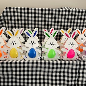May include: A row of eight white bunny-shaped Easter egg holders with colorful eggs inside. The holders are made of cardboard and have bunny ears, a face, and a tail. The eggs are in a rainbow of colors, including orange, yellow, green, blue, purple, and pink.