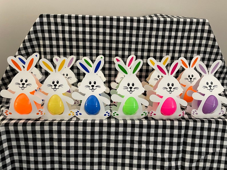 May include: A row of eight white bunny-shaped Easter egg holders with colorful eggs inside. The holders are made of cardboard and have bunny ears, a face, and a tail. The eggs are in a rainbow of colors, including orange, yellow, green, blue, purple, and pink.