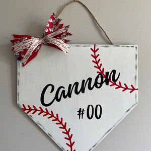 May include: White baseball home plate-shaped sign with red stitching and the name "Cannon" and the number "00" in black. A red and white bow with baseball-themed ribbon is attached to the top, along with a twine hanger.