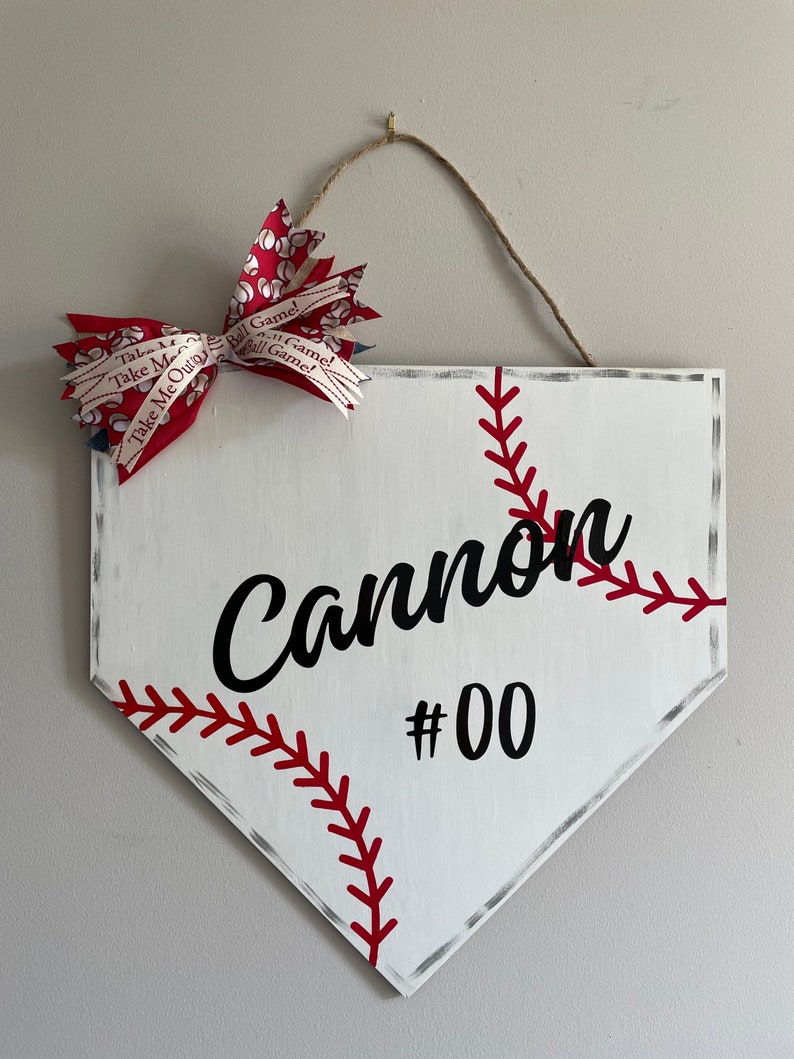 May include: White baseball home plate-shaped sign with red stitching and the name "Cannon" and the number "00" in black. A red and white bow with baseball-themed ribbon is attached to the top, along with a twine hanger.