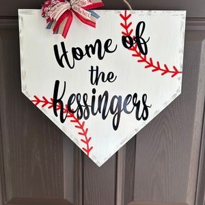 May include: White baseball home plate-shaped sign with red stitching and the text "Home of the Kessingers" in black script. A red, white, and blue bow is attached at the top with a jute rope for hanging. The sign is a decorative item.