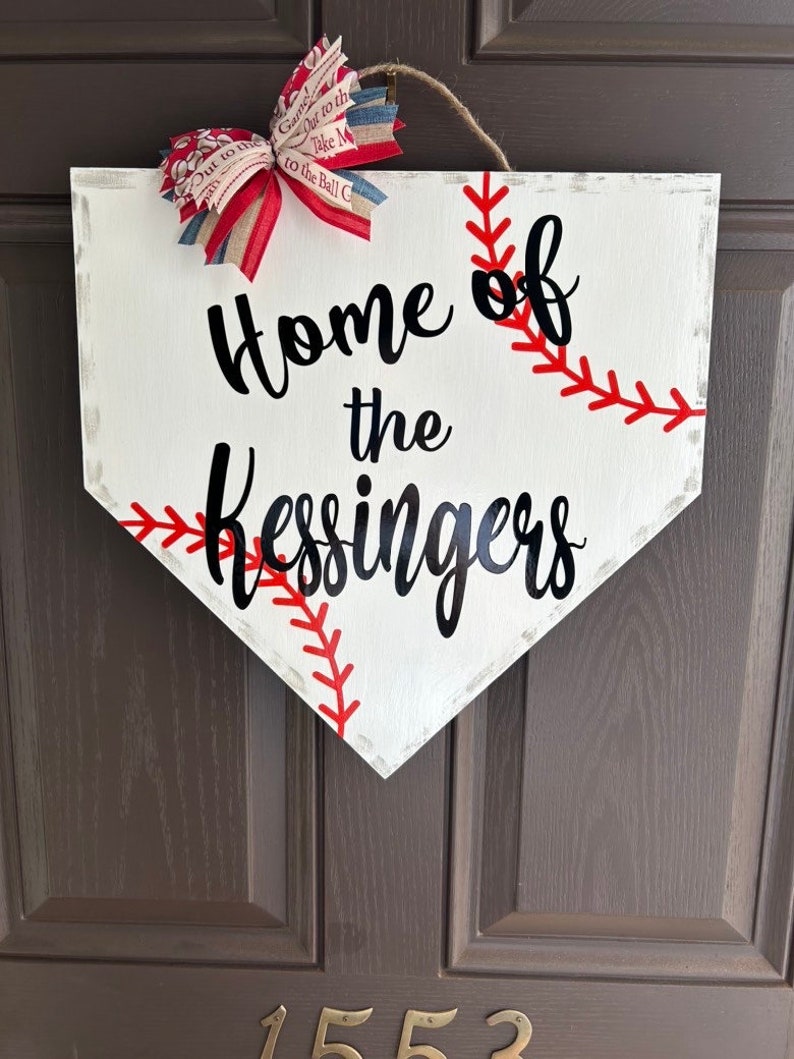 May include: White baseball home plate-shaped sign with red stitching and the text "Home of the Kessingers" in black script. A red, white, and blue bow is attached at the top with a jute rope for hanging. The sign is a decorative item.