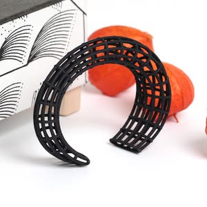 Könnte beinhalten: Ein schwarzes, geometrisches, durchbrochenes Armreif-Armband. Das Armband besteht aus einer Reihe von sich kreuzenden Linien, die ein gitterartiges Muster bilden.