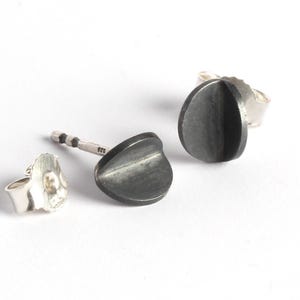 May include: Silver stud earrings with a dark grey, teardrop-shaped design. The earrings are displayed on a white surface, highlighting the contrasting colours and textures of the jewellery.