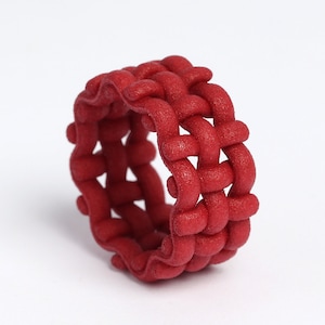 Op de afbeelding: Een rode geweven ring gemaakt van plastic. De ring is gemaakt van een reeks in elkaar grijpende lussen die een textuur creëren.