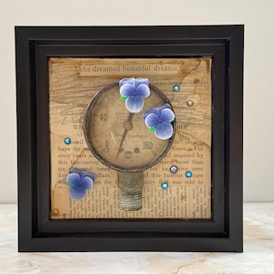 May include: Framed artwork featuring a vintage gauge with a cream-colored face and black numbers, overlaid on a collage of aged book pages. Adorned with three blue and purple flower embellishments and small blue gems. The frame is black.