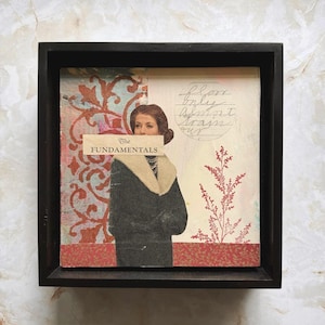 May include: Mixed media artwork in a black frame. The artwork features a vintage image of a woman in a black coat with a cream-colored collar. The text "The Fundamentals" is across the woman's face. A red floral design is on the left.