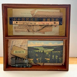 Könnte beinhalten: Eine zweistöckige Holzkiste mit Vintage-Ephemera. Der obere Bereich zeigt eine schwarze und braune elektrische Anschlussleiste und zerrissenes Papier. Der untere Bereich hat eine Miniatur-Eisenbahn und eine Landschaftspostkarte.