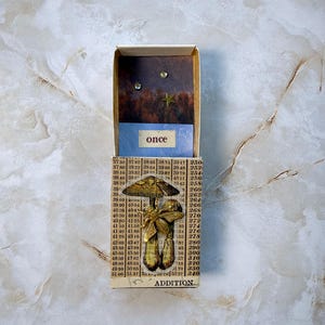 May include: A mixed media art piece featuring a small box with a miniature scene inside. The box contains a blue strip with the word "once" and a background with a star and two small gems. Below, a mushroom and bee-themed embellishment is attached.
