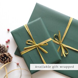 May include: Two green gift boxes wrapped with gold ribbon. The boxes are on a white surface with pine cones and red berries.