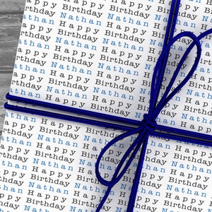May include: A white gift box wrapped in paper with the words "Happy Birthday Nathan" repeated in blue and black text. The box is tied with a blue ribbon.