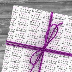 May include: A white gift box wrapped in paper with the words "Lucy Happy Birthday" repeated in pink and black. The box is tied with a purple ribbon.