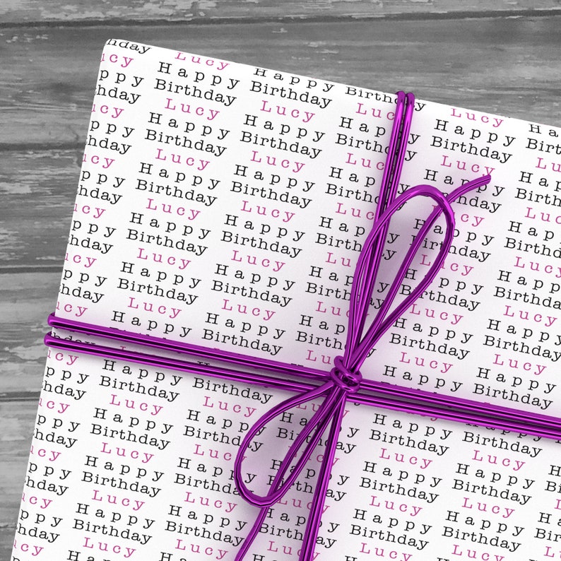 May include: A white gift box wrapped in paper with the words "Lucy Happy Birthday" repeated in pink and black. The box is tied with a purple ribbon.