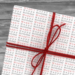 May include: A white gift box with red ribbon tied in a bow. The box is wrapped in paper with the words "Maggie Happy Birthday" repeated in red.