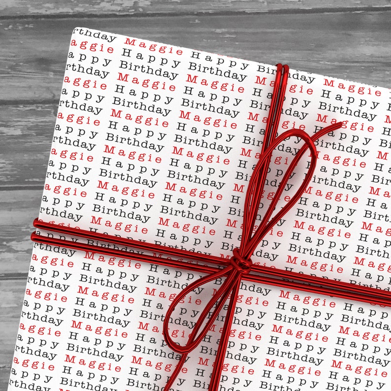 May include: A white gift box with red ribbon tied in a bow. The box is wrapped in paper with the words "Maggie Happy Birthday" repeated in red.