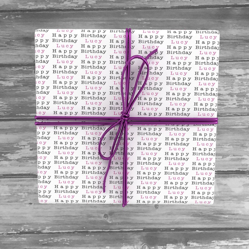 May include: A white gift box wrapped in pink and black patterned paper with the words "Lucy Happy Birthday" repeated. The box is tied with a purple ribbon.
