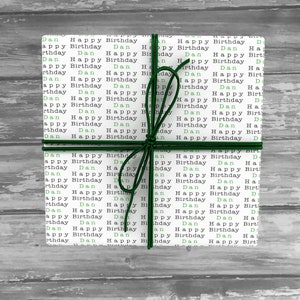 May include: A white gift box wrapped in paper with the words "Dan Happy Birthday" repeated in green and black. The box is tied with a green ribbon.
