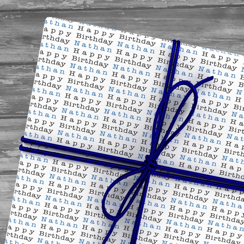 May include: A white gift box wrapped in paper with the words "Happy Birthday Nathan" repeated in blue and black text. The box is tied with a blue ribbon.