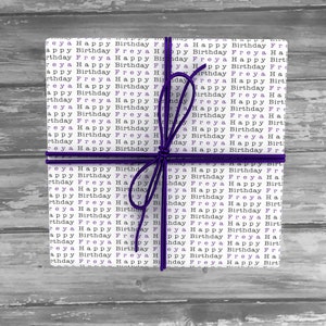May include: A white gift box wrapped in paper with the words "Happy Birthday Freya" repeated in black and purple. The box is tied with a purple ribbon.