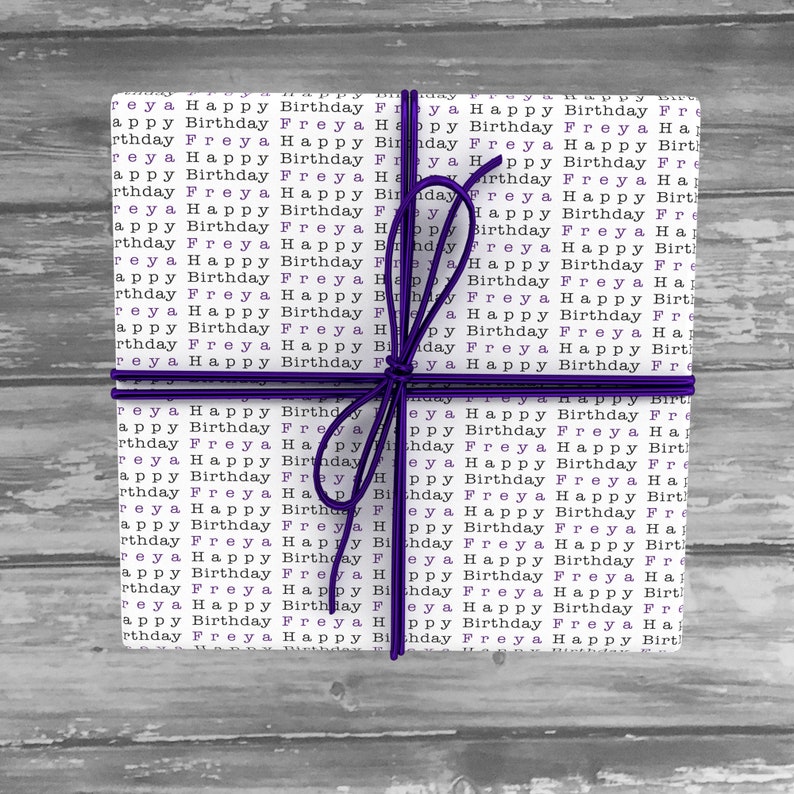 May include: A white gift box wrapped in paper with the words "Happy Birthday Freya" repeated in black and purple. The box is tied with a purple ribbon.