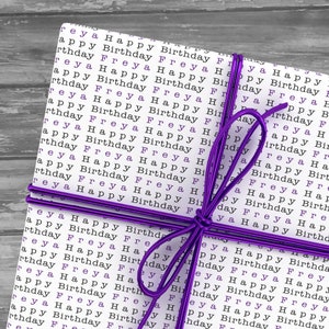 May include: A white gift box wrapped in paper with the words "Happy Birthday Freya" repeated in black text. The box is tied with a purple ribbon.