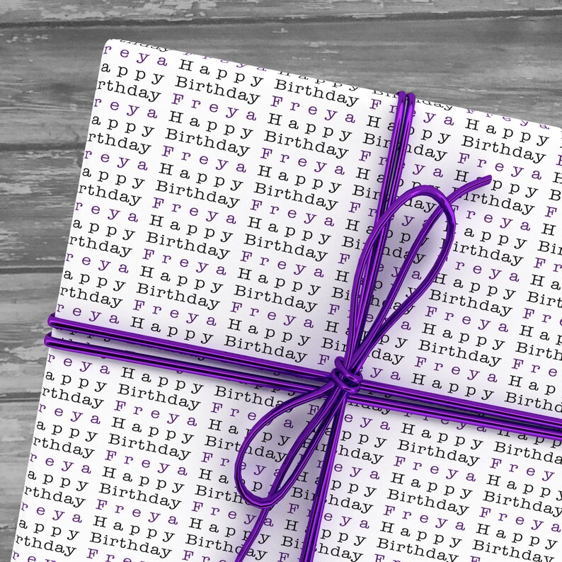 May include: A white gift box wrapped in paper with the words "Happy Birthday Freya" repeated in black text. The box is tied with a purple ribbon.