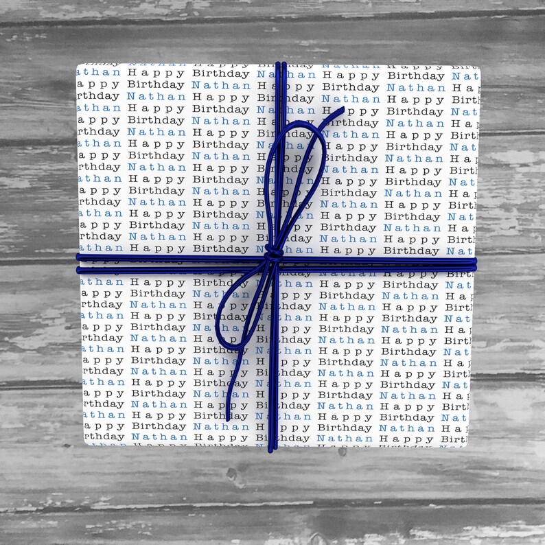 May include: A white gift box wrapped in blue and white paper with the words "Happy Birthday Nathan" repeated across the surface. The box is tied with a blue ribbon.
