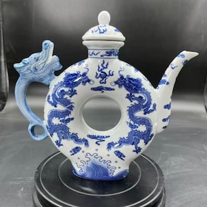 Puede incluir: Tetera de cerámica blanca con un diseño de dragón azul. La tetera tiene un cuerpo circular único con un pico y un asa en forma de dragón. La tapa está rematada con un pequeño pomo. El diseño presenta dragones azules y patrones de olas.