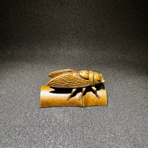 May include: A gold-colored cicada figurine perched on a small, cylindrical base. The cicada is intricately detailed with textured wings and legs.