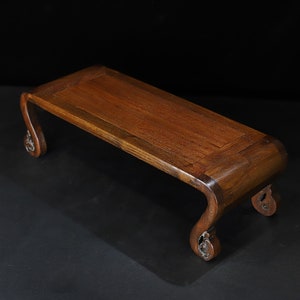 May include: A rectangular, wooden display stand with a rich, dark brown finish. The stand features curved legs with decorative carvings and a flat top surface, perfect for showcasing small items. The overall design is elegant and traditional.