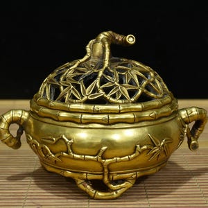 May include: A golden incense burner with a bamboo design. The lid features a bamboo leaf pattern and a curved handle. The body of the burner has bamboo-shaped handles and decorative bamboo stalks. The incense burner is made of metal.