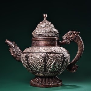 Könnte beinhalten: Eine Teekanne aus dunkler Bronze mit aufwendigen, geprägten Blumen- und Geometriemustern auf Körper und Deckel. Der Ausguss und der Griff sind drachenartig geformt, und der Fuß ist mit einem blütenblattartigen Design verziert, das detaillierte Metallarbeit zeigt.
