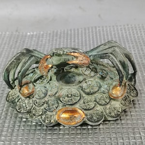 May include: A detailed bronze crab sculpture with copper-colored claws and accents. The crab sits atop a base adorned with coin-like details. The sculpture has a weathered, antique appearance.
