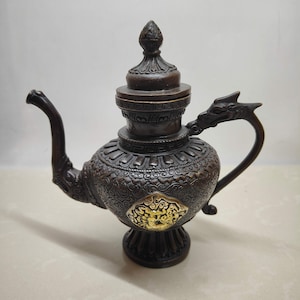 May include: A dark bronze-colored metal teapot featuring an intricate embossed pattern across its body, a dragon-shaped handle, and a pointed lid. A prominent, raised gold-colored circular medallion with detailed relief work is centered on the front.