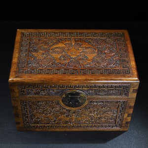Pode incluir: Uma caixa de madeira castanho escura com entalhes intrincados e uma trava de metal. A caixa tem um padrão geométrico na parte superior e nas laterais.